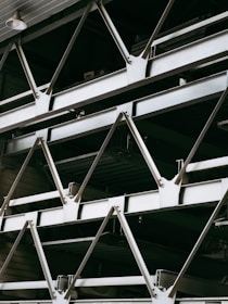 A close-up view of an industrial structure displaying several parallel metallic beams and cross-braces arranged in a geometric pattern. The beams are primarily composed of metal and exhibit a clean, angular design. A light fixture is visible at the top left corner.