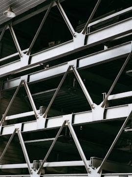 A close-up view of an industrial structure displaying several parallel metallic beams and cross-braces arranged in a geometric pattern. The beams are primarily composed of metal and exhibit a clean, angular design. A light fixture is visible at the top left corner.