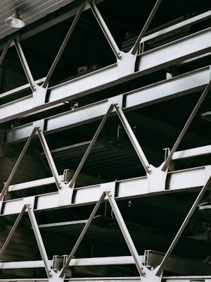 A close-up view of an industrial structure displaying several parallel metallic beams and cross-braces arranged in a geometric pattern. The beams are primarily composed of metal and exhibit a clean, angular design. A light fixture is visible at the top left corner.
