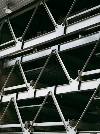 A close-up view of an industrial structure displaying several parallel metallic beams and cross-braces arranged in a geometric pattern. The beams are primarily composed of metal and exhibit a clean, angular design. A light fixture is visible at the top left corner.