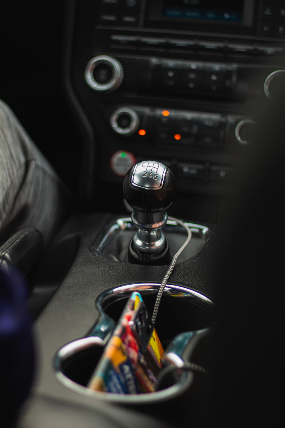 The image shows a car interior with various controls and features, including a silver gear shift on the center console, a radio dial, and a speedometer. The focus is on the gear shift, which has a black cover over it. A person's hand can be seen in the background, likely adjusting or interacting with one of these controls.