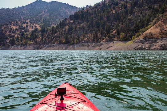 A red kayak is gliding across a large body of water surrounded by forested mountains. The water reflects sunlight, creating shimmering lines on its surface. The mountains are covered with dense greenery, and the sky is clear, suggesting a calm and serene environment.