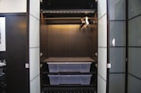 An empty wardrobe with open doors, featuring a dark interior. Inside, there are several metal bars for hanging clothes and two transparent plastic storage boxes on shelves. The wardrobe is sleek and modern, with a minimalist design.