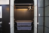 An empty wardrobe with open doors, featuring a dark interior. Inside, there are several metal bars for hanging clothes and two transparent plastic storage boxes on shelves. The wardrobe is sleek and modern, with a minimalist design.