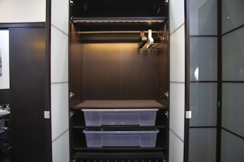 An empty wardrobe with open doors, featuring a dark interior. Inside, there are several metal bars for hanging clothes and two transparent plastic storage boxes on shelves. The wardrobe is sleek and modern, with a minimalist design.
