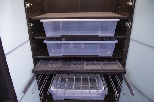 A dark-colored storage cabinet with two transparent plastic containers on separate shelves. Below them, there is a drawer with foam inserts, possibly for organization. The bottom section has metal rods, likely for hanging items, and another plastic container.