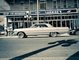 A vintage car is parked on a city street in front of a restaurant named Carmine's. The architecture features brick walls and large windows, with a distinctive sign promoting authenticity. Streetlights cast shadows, and a small group of people can be seen on the sidewalk.