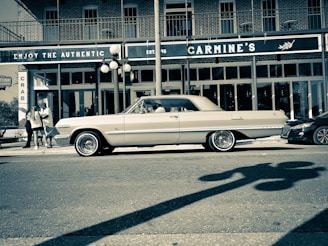A vintage car is parked on a city street in front of a restaurant named Carmine's. The architecture features brick walls and large windows, with a distinctive sign promoting authenticity. Streetlights cast shadows, and a small group of people can be seen on the sidewalk.