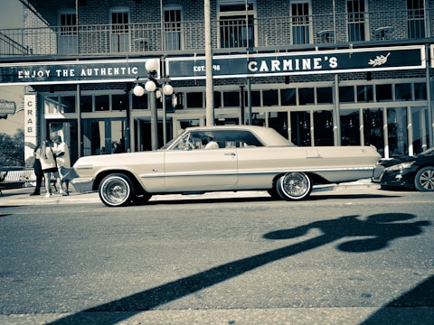 A vintage car is parked on a city street in front of a restaurant named Carmine's. The architecture features brick walls and large windows, with a distinctive sign promoting authenticity. Streetlights cast shadows, and a small group of people can be seen on the sidewalk.