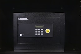 A black electronic safe with a keypad and a small digital display. The safe has a prominent brand logo on the front and a warning label at the top left corner. It is placed in a dark environment, enhancing its prominence and security features.
