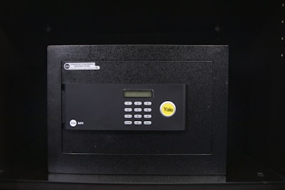 A black electronic safe with a keypad and a small digital display. The safe has a prominent brand logo on the front and a warning label at the top left corner. It is placed in a dark environment, enhancing its prominence and security features.
