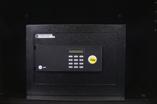 A black electronic safe with a keypad and a small digital display. The safe has a prominent brand logo on the front and a warning label at the top left corner. It is placed in a dark environment, enhancing its prominence and security features.