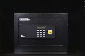 A black electronic safe with a keypad and a small digital display. The safe has a prominent brand logo on the front and a warning label at the top left corner. It is placed in a dark environment, enhancing its prominence and security features.