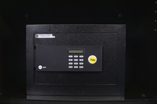 A black electronic safe with a keypad and a small digital display. The safe has a prominent brand logo on the front and a warning label at the top left corner. It is placed in a dark environment, enhancing its prominence and security features.