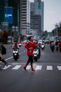 A person in a red athletic outfit is running across a zebra crossing on a busy urban street. They appear focused and energetic, holding a bottle in one hand. In the background, several motorcyclists wear helmets, and tall buildings line the street. The scene is set amidst a blend of natural and artificial lighting, indicating early morning or late afternoon.