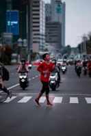 A person in a red athletic outfit is running across a zebra crossing on a busy urban street. They appear focused and energetic, holding a bottle in one hand. In the background, several motorcyclists wear helmets, and tall buildings line the street. The scene is set amidst a blend of natural and artificial lighting, indicating early morning or late afternoon.