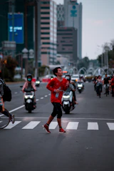 A person in a red athletic outfit is running across a zebra crossing on a busy urban street. They appear focused and energetic, holding a bottle in one hand. In the background, several motorcyclists wear helmets, and tall buildings line the street. The scene is set amidst a blend of natural and artificial lighting, indicating early morning or late afternoon.