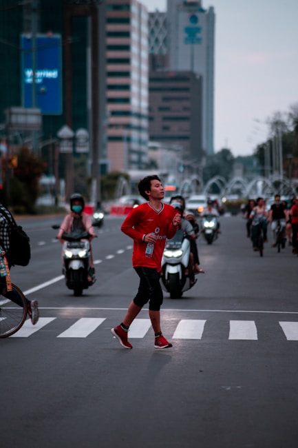 A person in a red athletic outfit is running across a zebra crossing on a busy urban street. They appear focused and energetic, holding a bottle in one hand. In the background, several motorcyclists wear helmets, and tall buildings line the street. The scene is set amidst a blend of natural and artificial lighting, indicating early morning or late afternoon.