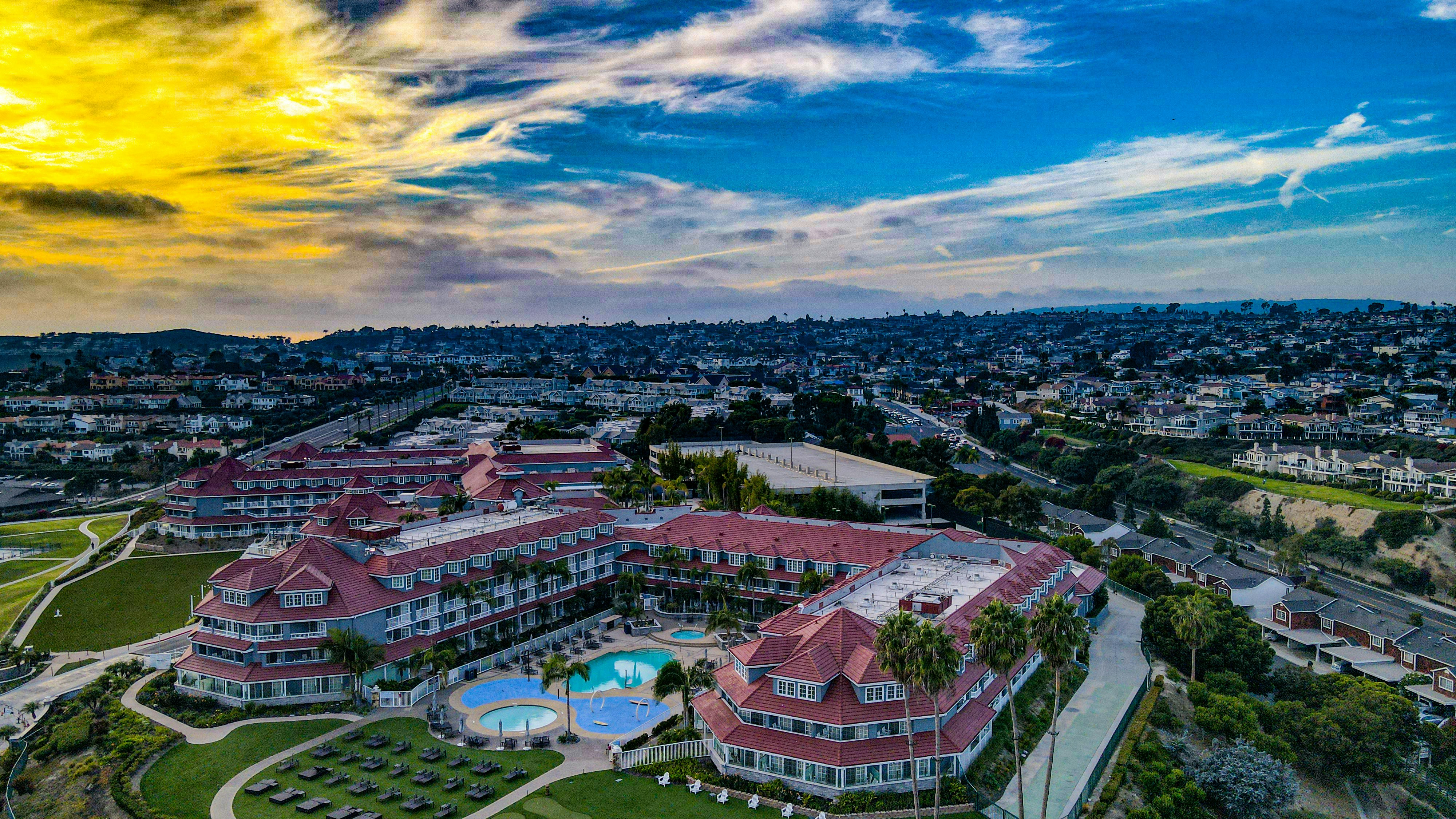 A large residential or hotel complex with red-roofed buildings is surrounded by lush green lawns and a swimming pool area. The complex is located on a hillside with a view of a densely packed urban landscape in the background. The sky is a vibrant mix of yellow and blue, with scattered clouds.