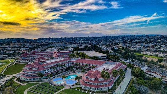 A large residential or hotel complex with red-roofed buildings is surrounded by lush green lawns and a swimming pool area. The complex is located on a hillside with a view of a densely packed urban landscape in the background. The sky is a vibrant mix of yellow and blue, with scattered clouds.