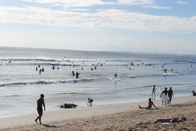 A sunny day at the beach with people enjoying various activities. Numerous surfers ride the waves in the ocean, while others relax on the sandy shore. The ocean stretches out into the horizon under a partly cloudy sky.