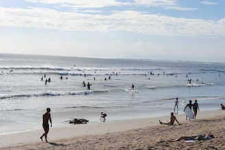 A sunny day at the beach with people enjoying various activities. Numerous surfers ride the waves in the ocean, while others relax on the sandy shore. The ocean stretches out into the horizon under a partly cloudy sky.