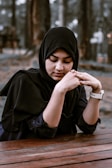 A woman wearing a black hijab is sitting at a wooden table with her hands gently clasped. The background is blurred with trees, suggesting an outdoor setting.