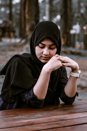 A woman wearing a black hijab is sitting at a wooden table with her hands gently clasped. The background is blurred with trees, suggesting an outdoor setting.