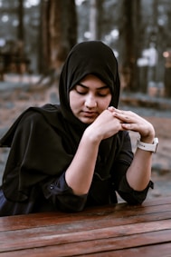 A woman wearing a black hijab is sitting at a wooden table with her hands gently clasped. The background is blurred with trees, suggesting an outdoor setting.