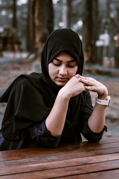 A woman wearing a black hijab is sitting at a wooden table with her hands gently clasped. The background is blurred with trees, suggesting an outdoor setting.