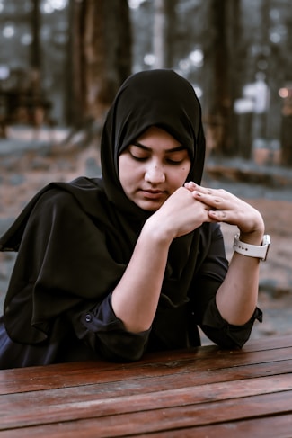A woman wearing a black hijab is sitting at a wooden table with her hands gently clasped. The background is blurred with trees, suggesting an outdoor setting.