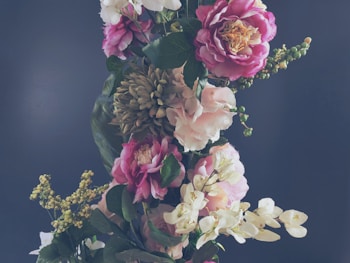 An elegant arrangement of various flowers, including pink roses and white orchids, set against a dark background. The flowers are surrounded by lush green leaves and some small budding green plants.