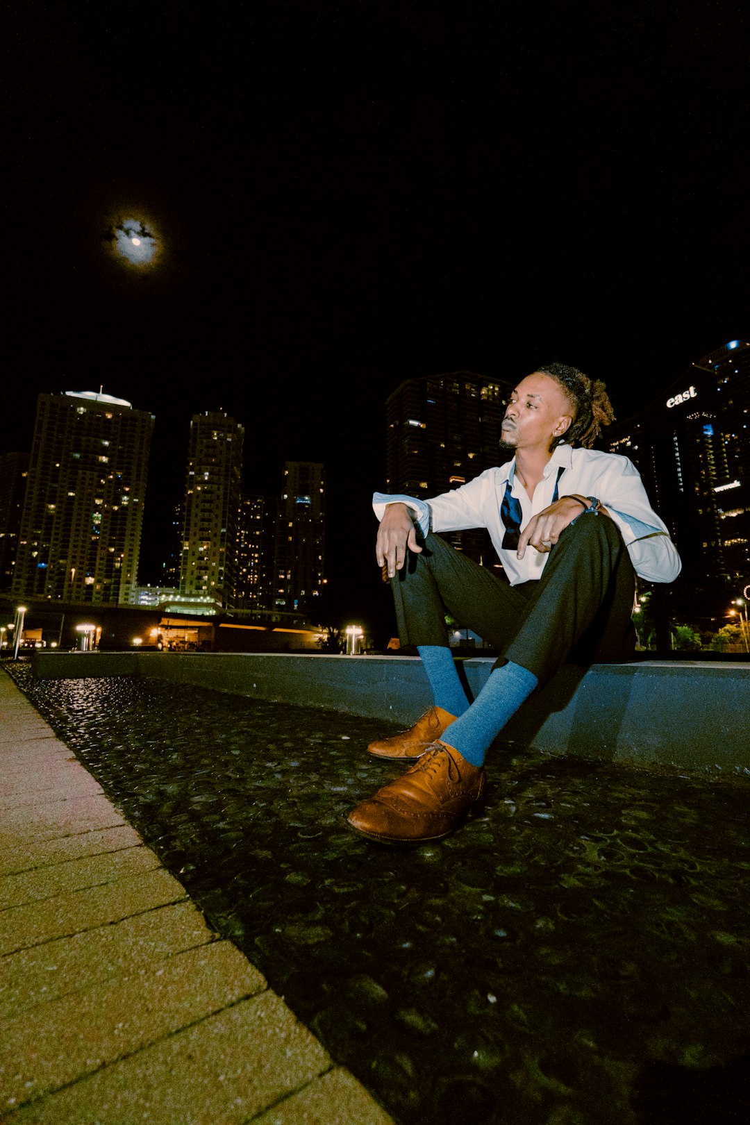 man in white dress shirt and black pants sitting on gray concrete pavement during night time