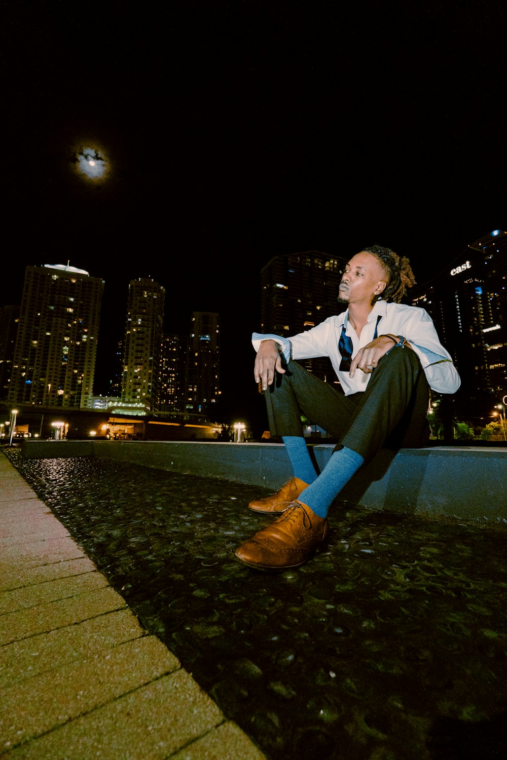 Mann in weißem Hemd und schwarzer Hose sitzt nachts auf grauem Betonpflaster