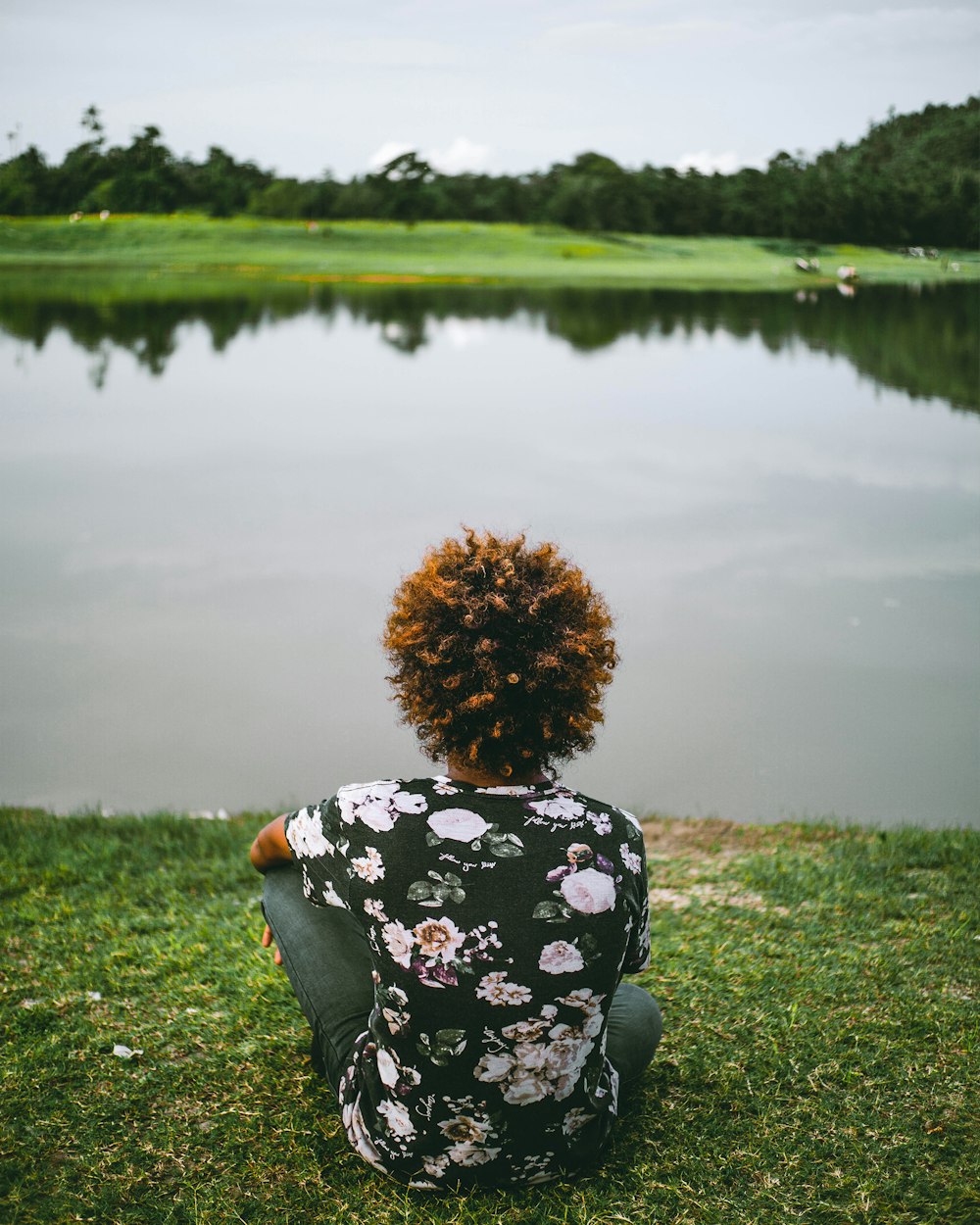 woman in black and white floral long sleeve shirt sitting on green grass field near lake