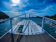 A wooden boat deck is surrounded by a netted railing, with a view of a serene body of water and distant green hills under a bright blue sky. The sunlight is partially hidden by clouds, casting reflections on the water.