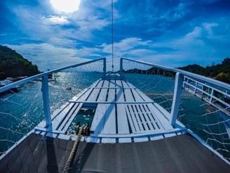 A wooden boat deck is surrounded by a netted railing, with a view of a serene body of water and distant green hills under a bright blue sky. The sunlight is partially hidden by clouds, casting reflections on the water.