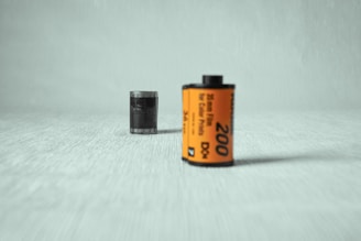A 35mm film roll with an orange label stands on a textured, light-colored surface. The film is prominently featured in the foreground, displaying the number 200 and text indicating it is for color prints. The background is softly blurred, focusing attention on the film roll.