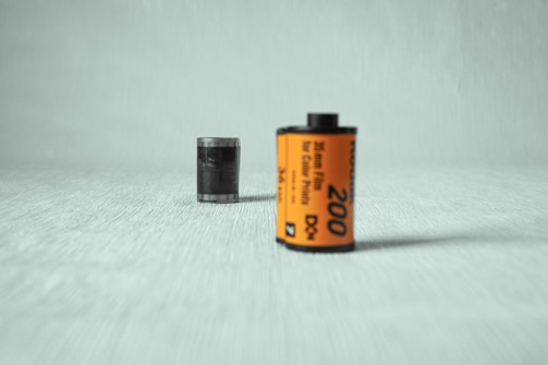 A 35mm film roll with an orange label stands on a textured, light-colored surface. The film is prominently featured in the foreground, displaying the number 200 and text indicating it is for color prints. The background is softly blurred, focusing attention on the film roll.