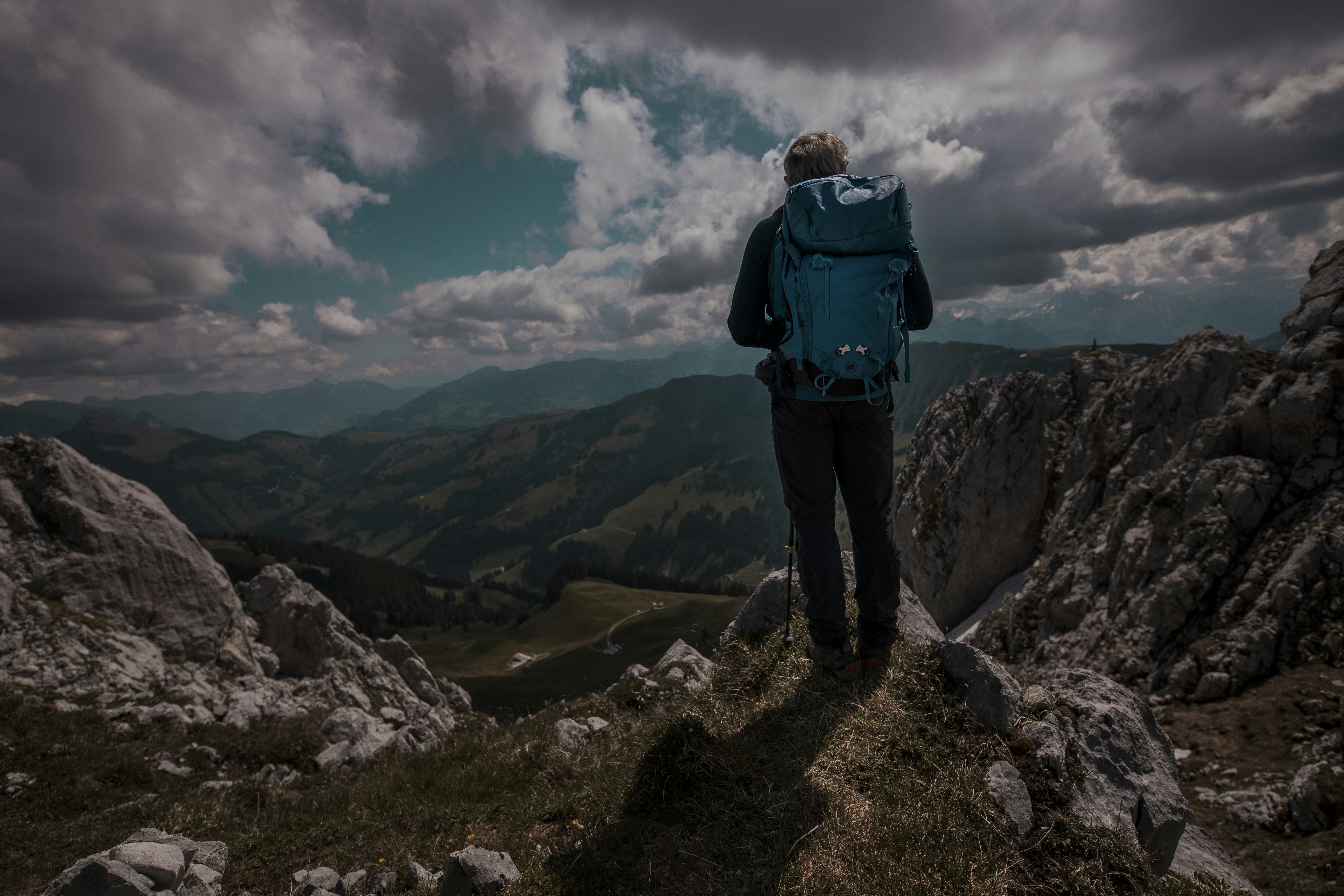man in blue jacket and black pants standing on rocky mountain under white clouds during daytime