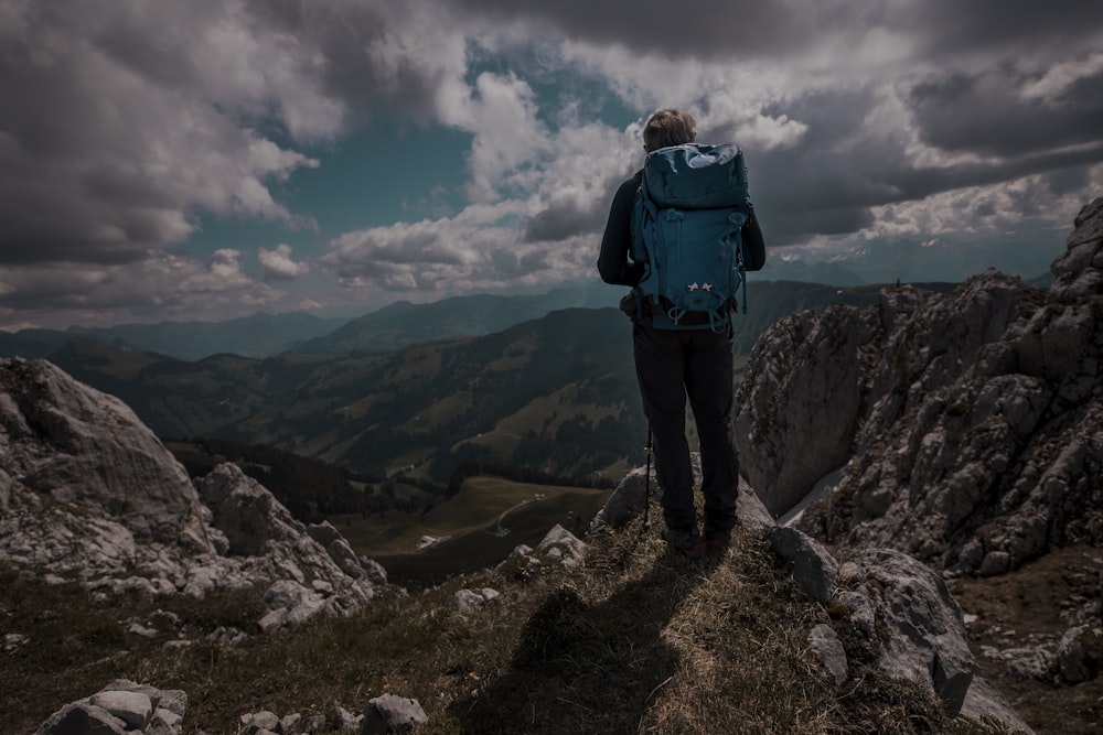 man in blue jacket and black pants standing on rocky mountain under white clouds during daytime