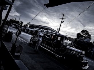 A dark and moody street scene depicts a traditional jeepney parked at a curb under a cloudy sky. Power lines crisscross the frame, adding to the urban feel. A person wearing a hat is standing beside the jeepney.