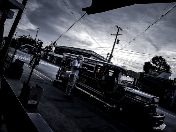 A dark and moody street scene depicts a traditional jeepney parked at a curb under a cloudy sky. Power lines crisscross the frame, adding to the urban feel. A person wearing a hat is standing beside the jeepney.