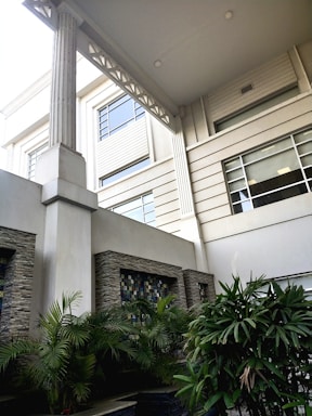 A modern architectural courtyard featuring large pillars, stone walls with decorative tiles, and lush green plants. The upper level showcases horizontal windows and clean, geometric lines.