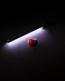A red light bulb lies on a dark surface beside a strip of bright white LED lights. The LED lights cast a soft glow around them, creating a contrast between the bright light and the darkness of the background.