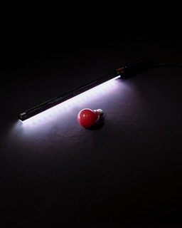 A red light bulb lies on a dark surface beside a strip of bright white LED lights. The LED lights cast a soft glow around them, creating a contrast between the bright light and the darkness of the background.