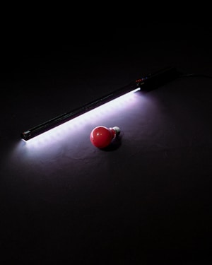 A red light bulb lies on a dark surface beside a strip of bright white LED lights. The LED lights cast a soft glow around them, creating a contrast between the bright light and the darkness of the background.