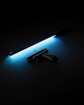 A handgun is placed beside a long horizontal strip of blue neon light against a dark background, creating a stark contrast and highlighting the silhouette of the weapon.