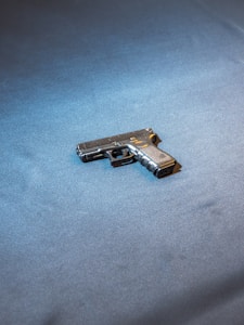 A handgun rests on a flat, blue-gray surface with soft lighting highlighting its metallic features. The weapon is centered in the image, with shadows giving it a three-dimensional appearance.