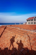 A rooftop with red shingles casting the shadow of a Ferris wheel in the foreground. A building with a gable roof stands to the right, with windows reflecting daylight. The background features a clear blue sky and a distant view of the sea.
